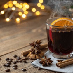 Tasse de café garnie d'une tranche d'orange séchée, posée sur serviette grise avec bâtons de cannelle, étoiles d'anis, grains de café et guirlande lumineuse dorée floue en arrière-plan.