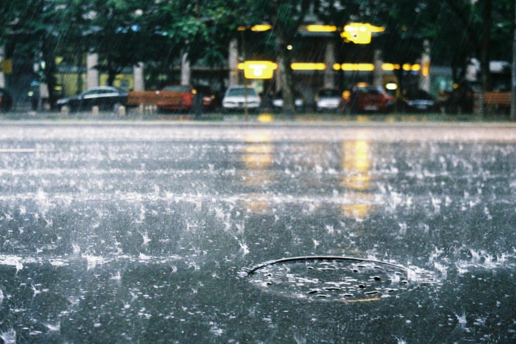 Cafe938_MoutureMeteo_Pluie » Café 9|38 Rainy street scene with rain splashes on the camera lens, blurred cars parked along a street, and yellow station-style lights in the background reflecting off the wet pavement.