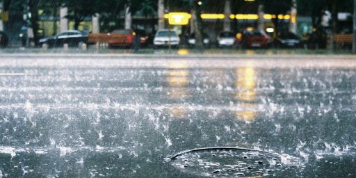 Rainy street scene with rain splashes on the camera lens, blurred cars parked along a street, and yellow station-style lights in the background reflecting off the wet pavement.