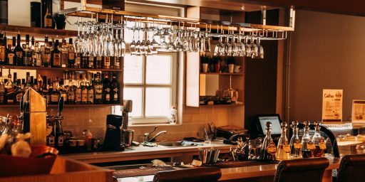 Intérieur chaleureux d'un bar professionnel avec comptoir en bois, étagères garnies de bouteilles de spiritueux, verres suspendus au plafond, machine à espresso visible et éclairage naturel provenant d'une fenêtre. L'espace évoque l'atmosphère sophistiquée d'un établissement spécialisé dans la mixologie de qualité.