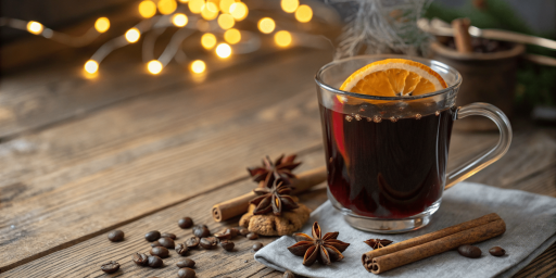 Tasse de café garnie d'une tranche d'orange séchée, posée sur serviette grise avec bâtons de cannelle, étoiles d'anis, grains de café et guirlande lumineuse dorée floue en arrière-plan.